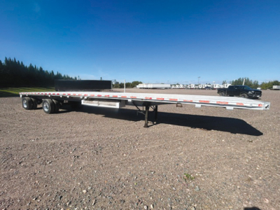 Flatbed trailer with aluminum deck showing long low profile and support legs, side rails with reflective tape, dual rear axles, and a gooseneck-capable front area visible.