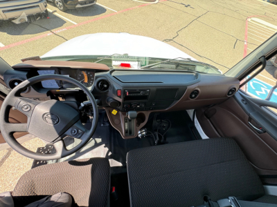 Truck cab interior of a 2025 Hino L6 showing steering wheel, dashboard, gear selector, and front seats from the driver’s perspective.