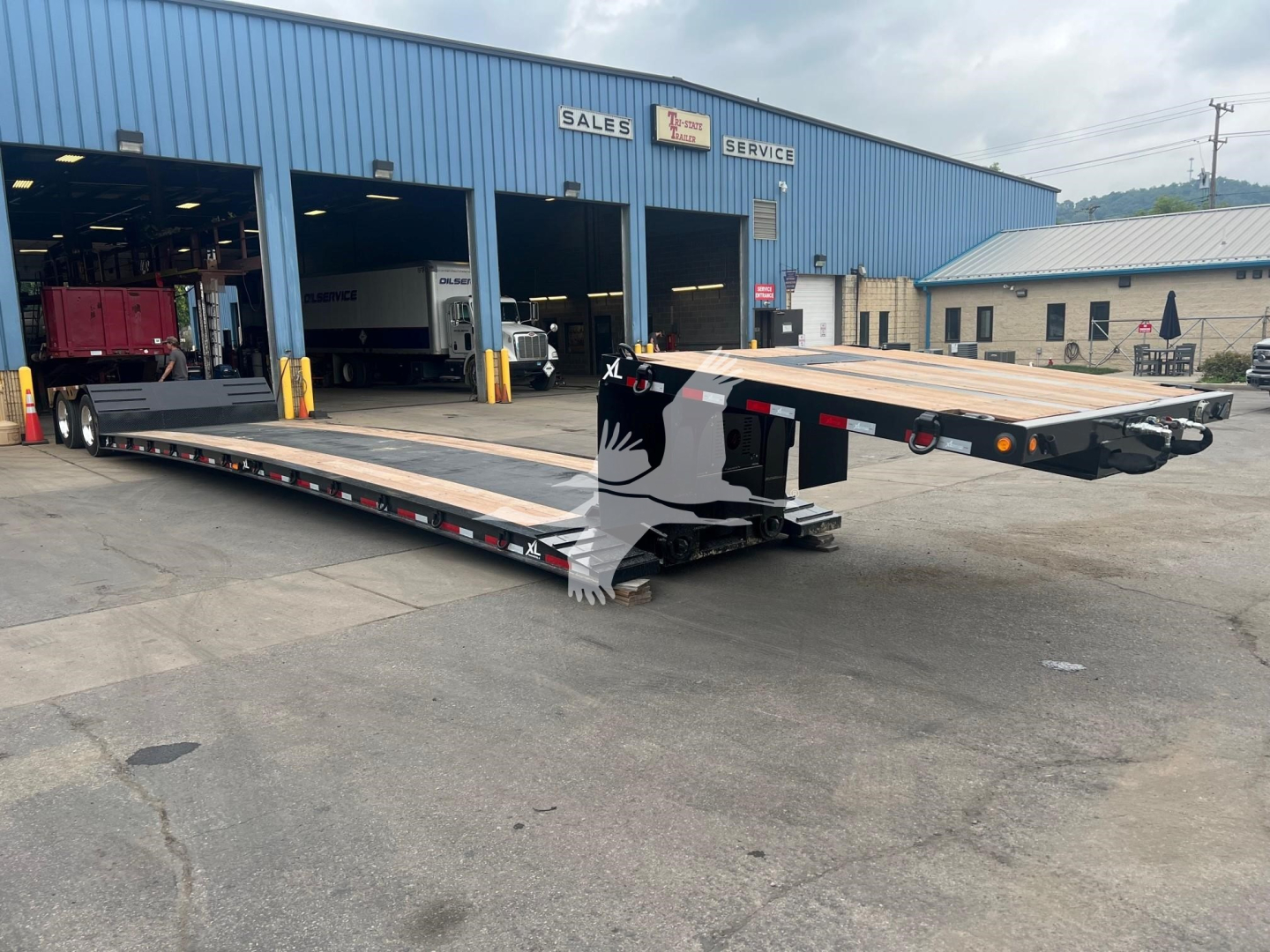 XL Specialized mini deck trailer in service bay showing a long low-profile flatbed with a dropped gooseneck, black frame, wooden deck, and safety decals at rear.