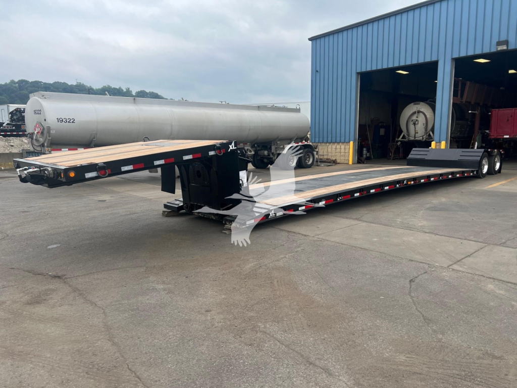 Trailer showing XL Specialized mini deck with long flatbed and dual drop expandable trailer in salvage yard. Tanker trailer in background with blue building.