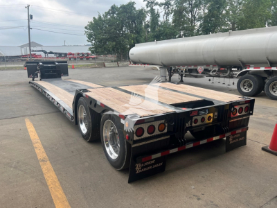 Trailer showing a flatbed lowboy with wooden deck, dual rear axles, and mud flaps; visible are tail lights, rear bumper, and reflective striping. No clear text readable on components.
