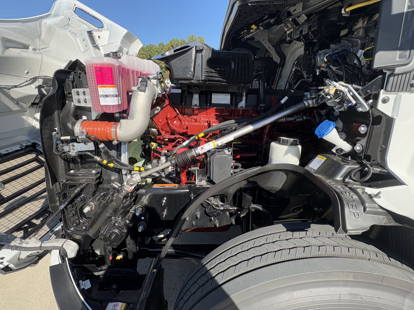 Engine bay of a Mack PR64T truck showing red engine, pink coolant reservoir, exposed hoses, and surrounding components.