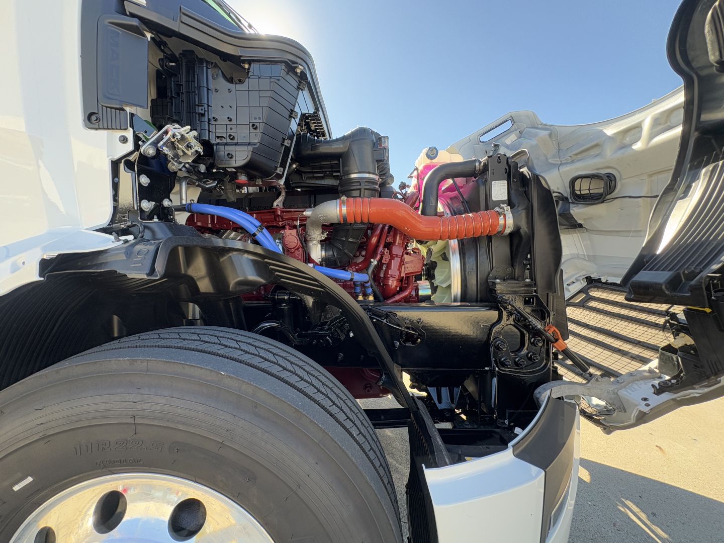 Front-side engine bay of a 2026 Mack PR64T conventional daycab showing red engine, orange coolant hose, blue wiring, and black chassis with wheel in foreground.