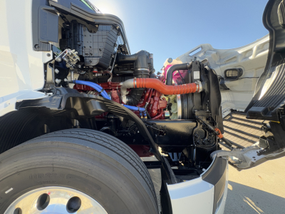 Front-side engine bay of a 2026 Mack PR64T conventional daycab showing red engine, orange coolant hose, blue wiring, and black chassis with wheel in foreground.