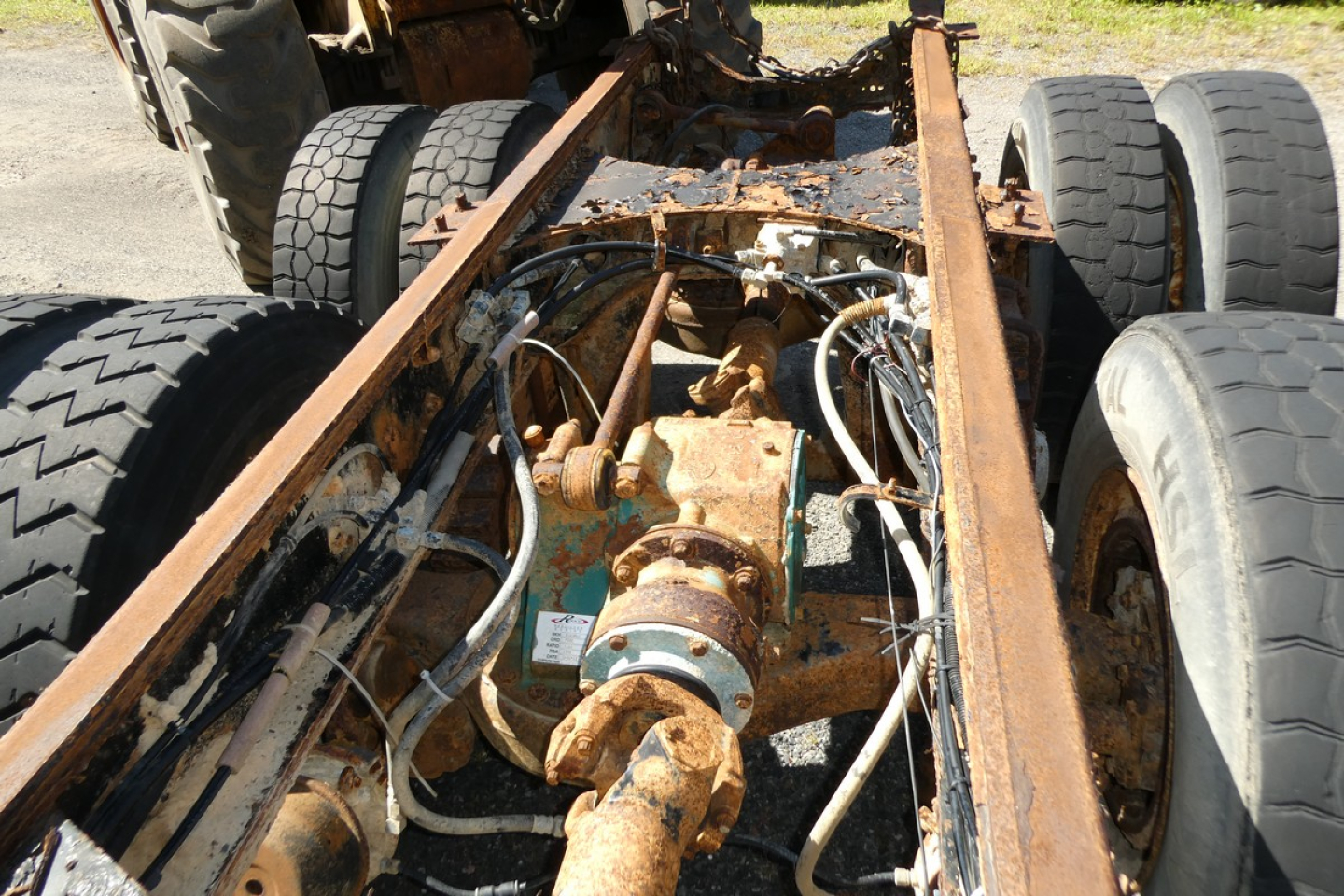 Vehicle frame and drivetrain coupling with steering shaft and hoses in view; rusty metal chassis and dual rear wheels under a cab and chassis truck.