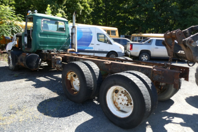 Mack RD688S cab chassis with exposed frame and dual rear axles, rusted wheels, and nearby vehicles in a yard.