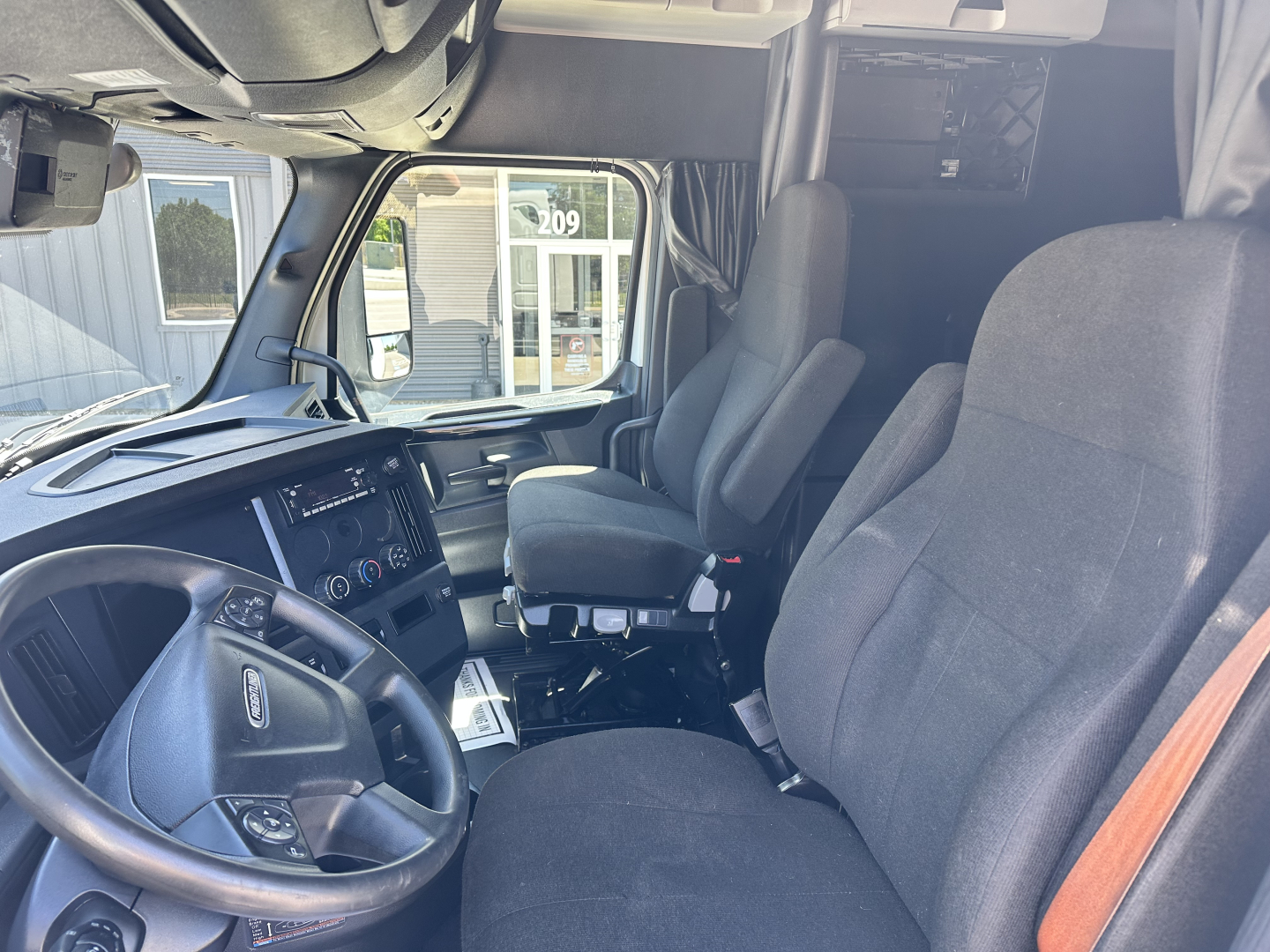Truck cab interior of a 2023 Freightliner Cascadia 126, driver seat and passenger seat visible, dashboard with controls, steering wheel, and side window.