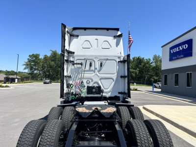 Freightliner Cascadia 126 truck rear view showing chassis with dual rear axles and fifth wheel, empty sleeper cab interior, and a building and flag in background.