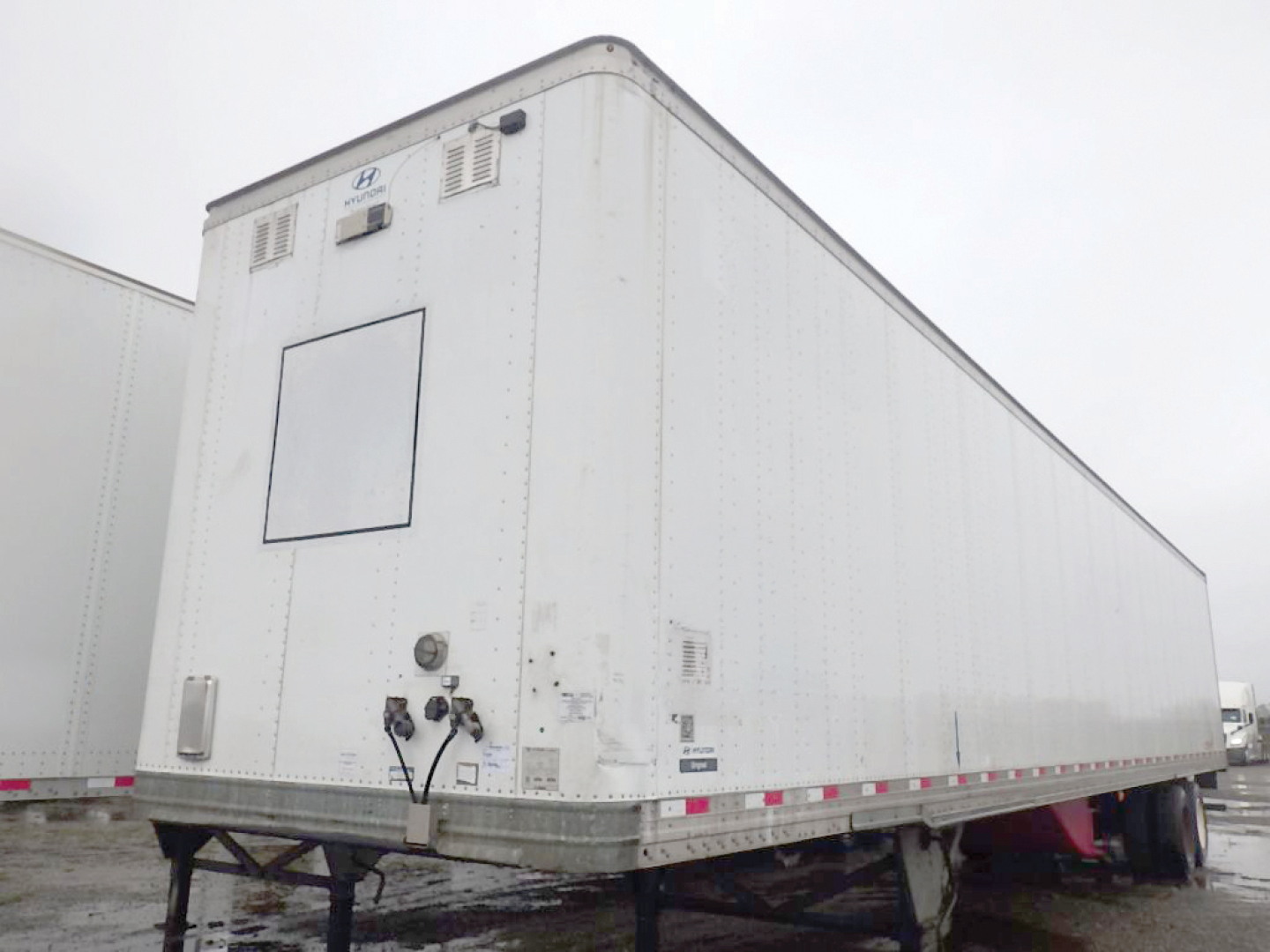 Trailer rear view showing mounted Hyundai logo, plumbing and utility panels, with doors and light fixtures on a white cargo trailer.