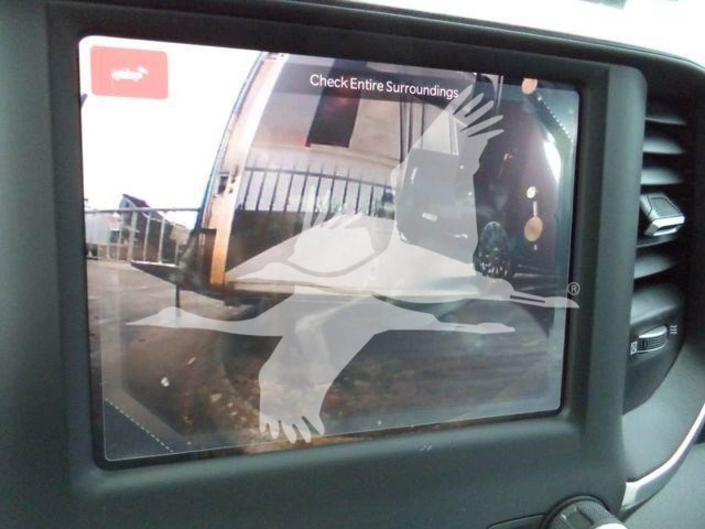 In-vehicle display showing a rearview camera image with a silhouette graphic and text 'Check Entire Surroundings' at the top.