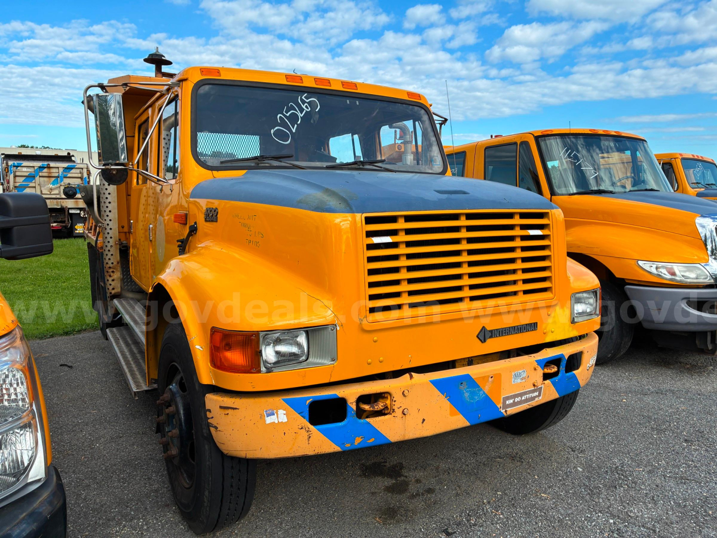 Front view of an orange 2002 International 4700 dump truck with a black grille and blue diagonal stripes on the bumper, parked among other orange trucks.
