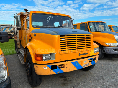 Front view of an orange 2002 International 4700 dump truck with a black grille and blue diagonal stripes on the bumper, parked among other orange trucks.