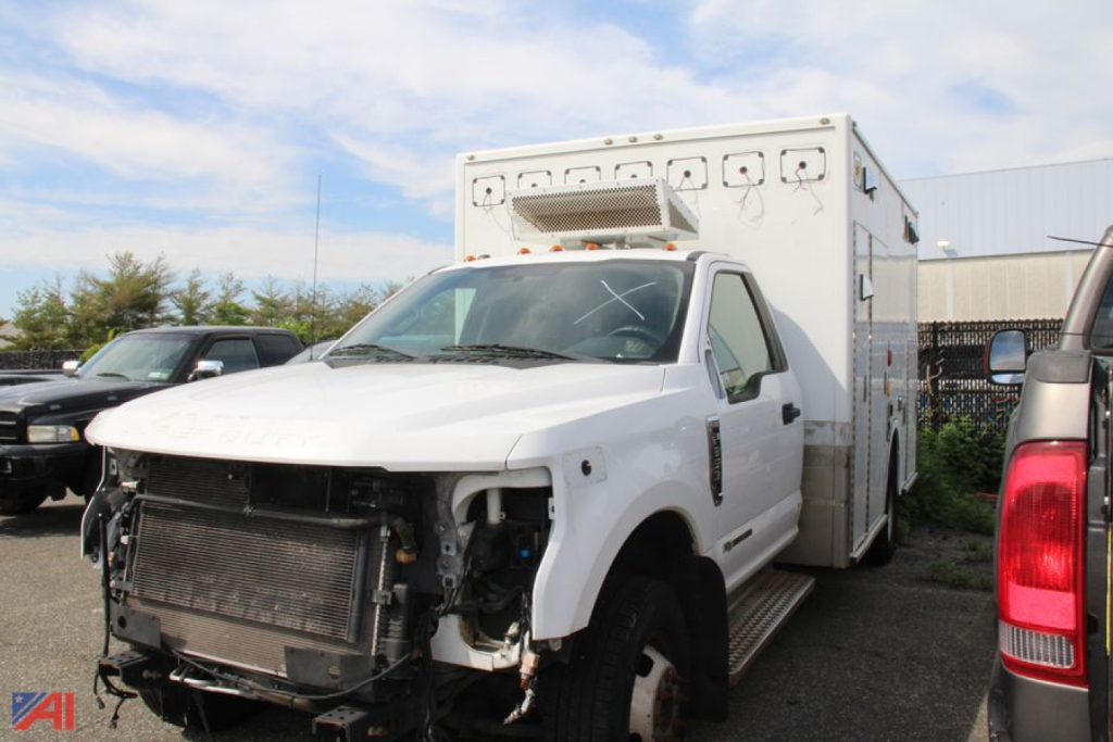Ambulance chassis with front bumper and grille removed, white cab, large box rear, equipment compartments, Ford F-350 Super Duty ambulance in serviceable condition.