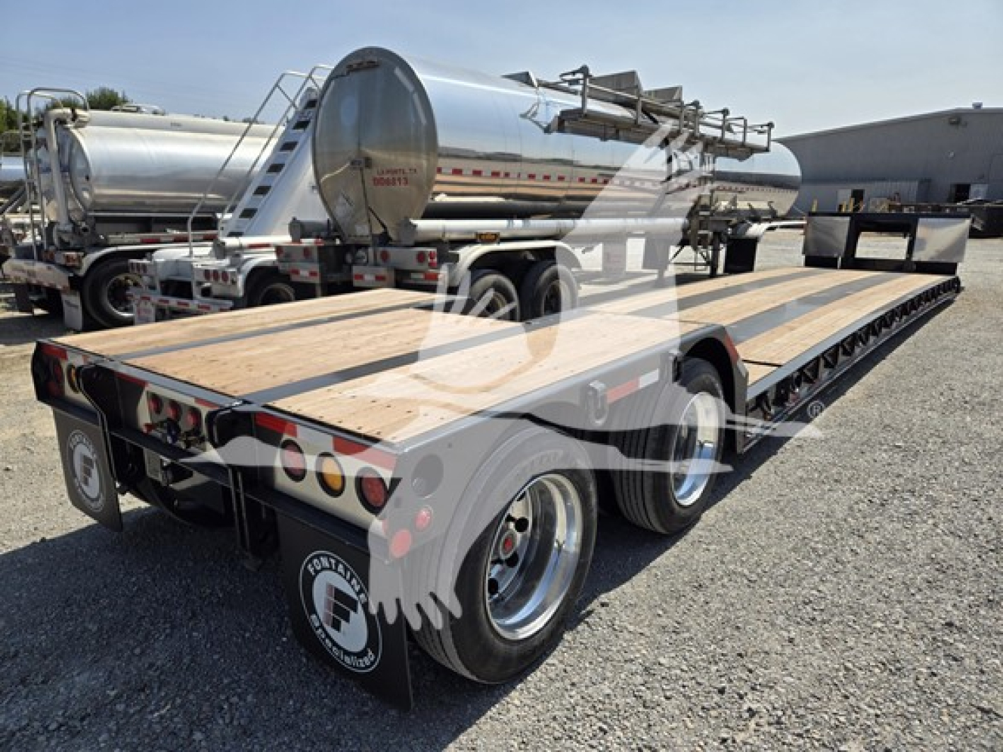 Flatbed trailer with triple-axle configuration and tanker trailer in background, viewed from rear-left, brand labels not legible.