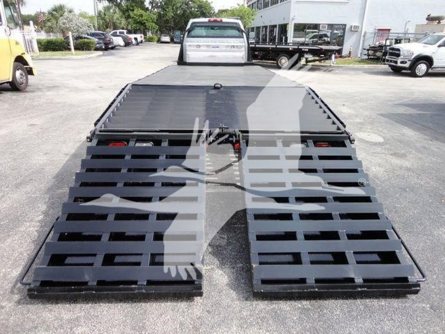 Flatbed truck bed of a 2023 Chevrolet Silverado 6500HD with two hinged ramps extended, black metal slats, rear view with surrounding lot and other trucks.