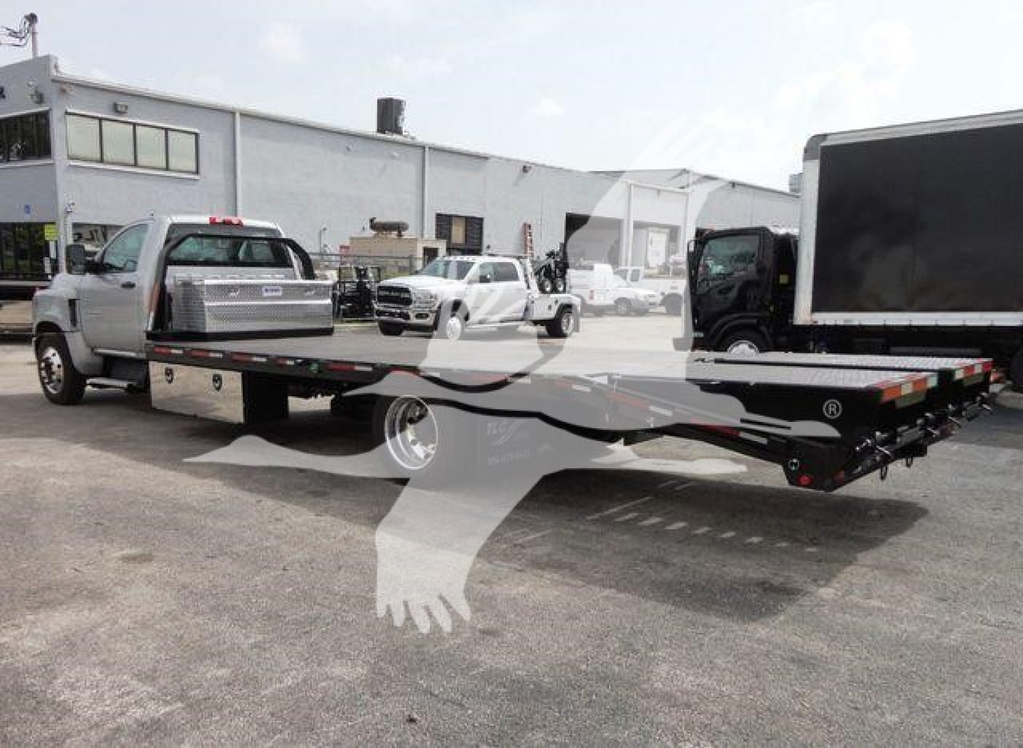Flatbed tow truck trailer connected to a Chevrolet Silverado 6500HD in a dealership lot. Text visible on truck includes no discernible model badges or stickers due to watermark.