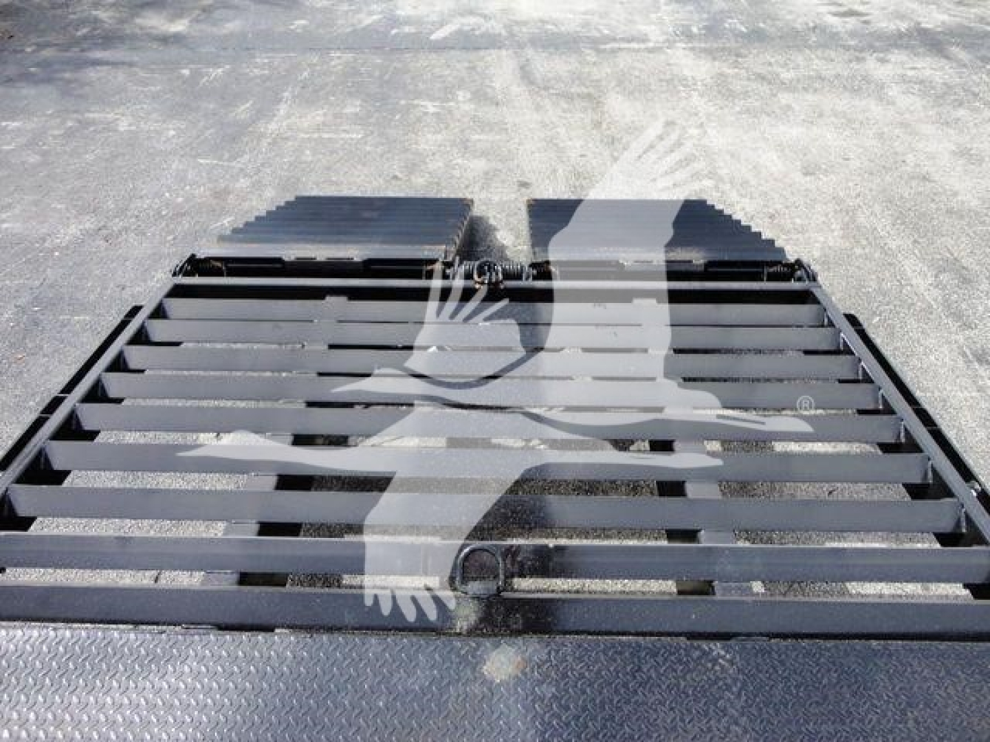 Flatbed truck bed with a black grated surface and metal ramp sections. Logo watermark across center. Visible wear and distant asphalt.