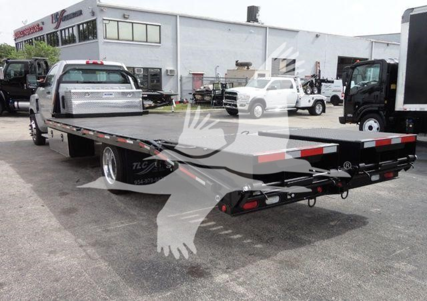 Flatbed tow truck trailer with dual rear ramps and black bed, parked in a lot with other trucks; side rails and reflective tape visible.
