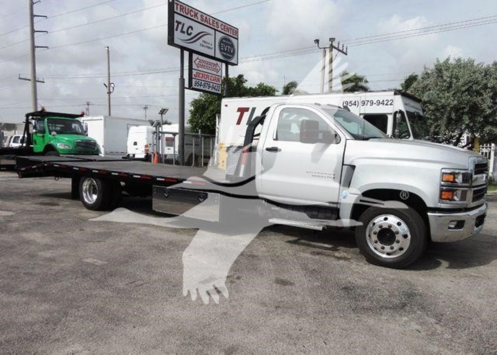 Flatbed tow truck with white cab and long black flatbed, Chevrolet Silverado 6500HD branding not visible from side; dealership lot in background.