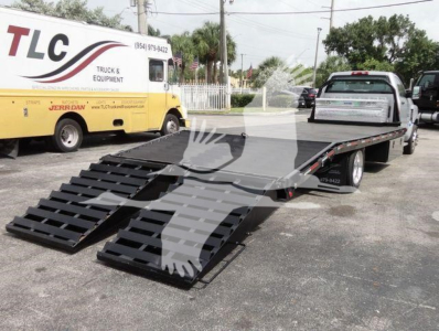Flatbed tow truck with dual ramps extending from the back. Chevrolet Silverado 6500HD flatbed with ramp system visible; TLC Trucks & Equipment signage on yellow truck in background.