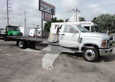 Flatbed tow truck with white cab and long black flatbed, Chevrolet Silverado 6500HD branding not visible from side; dealership lot in background.