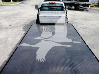 Flatbed tow truck bed with diamond plate steel surface and a white Chevrolet Silverado 6500HD cab in the background; emblem and toolbox visible on the front of the bed.