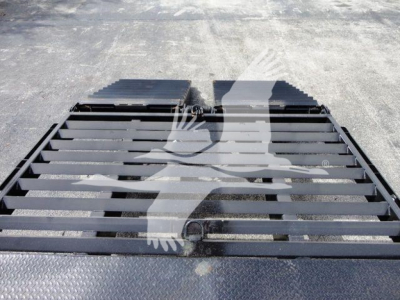 Flatbed truck bed with a black grated surface and metal ramp sections. Logo watermark across center. Visible wear and distant asphalt.