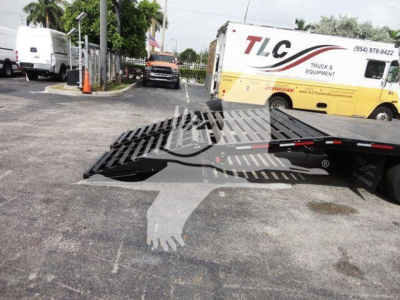 Flatbed tow truck with a folded metal ramp in the foreground, TLC Truck & Equipment van in background, parking lot scene