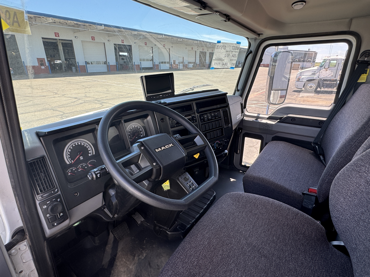 Mack MD7 cab interior showing steering wheel, dashboard gauges, and center infotainment screen; driver seat and passenger seat visible; exterior lot with a building in background.