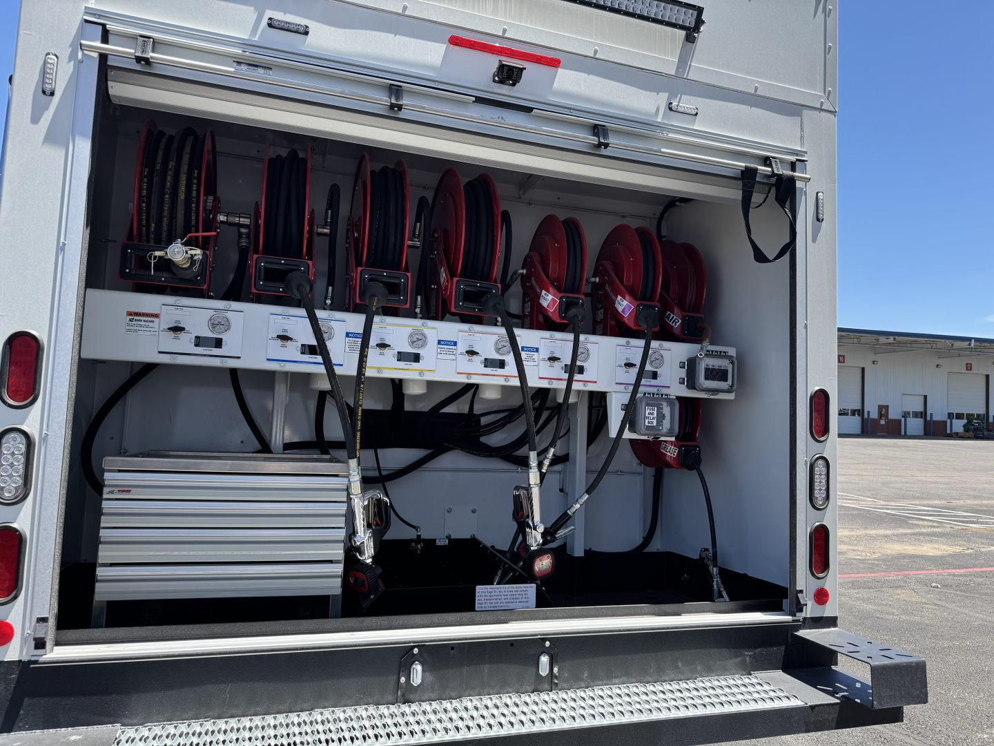 Back open service body showing reel hoses and equipment storage on a Mack MD7 chassis. Visible are red hose reels mounted on a white interior with electrical and control panels and a silver toolbox.