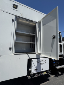 Service body door open showing interior shelves and exterior equipment compartment with label 'RANAIR', 'PNE', and electrical box. Vehicle side panel with storage cabinet.