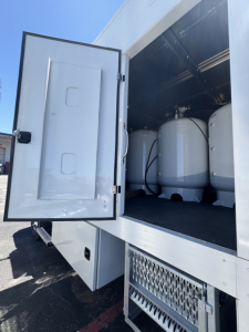 Truck rear showing storage with large insulated tanks; open external storage compartment on a new 2025 Mack MD7 service/utility truck; shelves and metal steps visible.