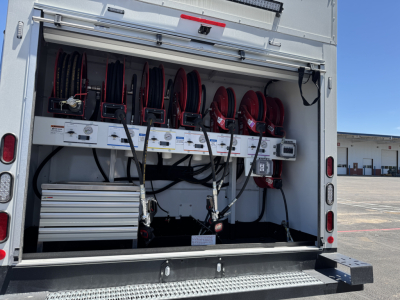 Back open service body showing reel hoses and equipment storage on a Mack MD7 chassis. Visible are red hose reels mounted on a white interior with electrical and control panels and a silver toolbox.