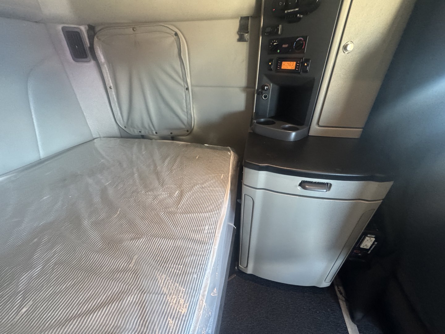 Interior sleeper area of a 2023 Peterbilt 579 showing bed with gray sheet, overhead storage, and control panel with radio in center console.