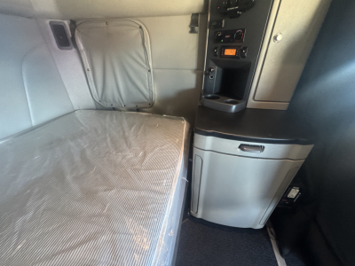 Interior sleeper area of a 2023 Peterbilt 579 showing bed with gray sheet, overhead storage, and control panel with radio in center console.