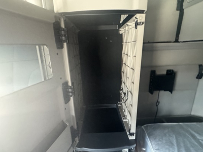 Cab interior view of a Peterbilt 579 sleeper cab showing a small storage compartment with a hinged door, metal ladder rungs, and a dark open space beyond.