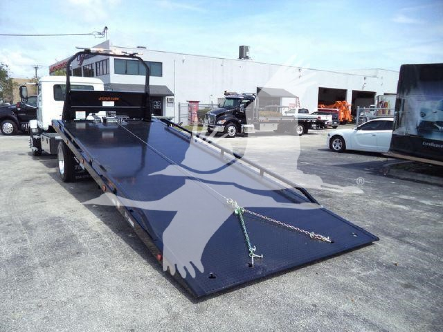 Flatbed rollback tow truck from left side, long black steel deck with ramp down, chains secured at the ramp tip, Mack Md6 listing details in image context.