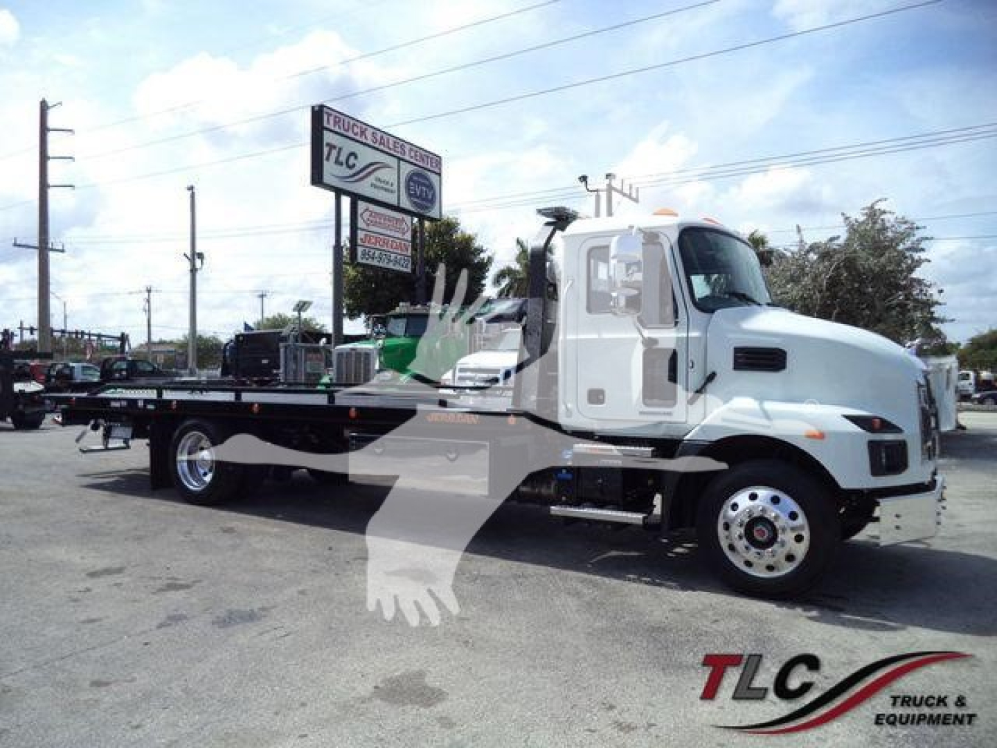 Flatbed tow truck showing new 2027 Mack MD6 with rollback equipment and dealer lot in background.