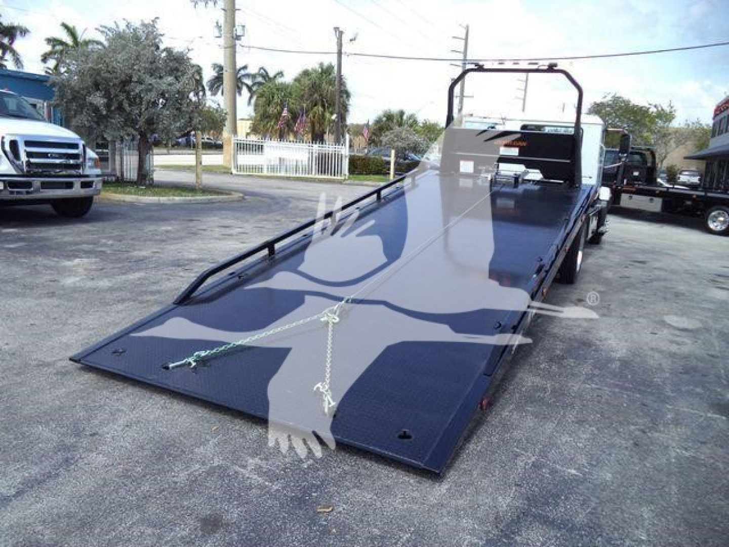 Flatbed rollback truck in a parking lot with a lowered, black metal ramp and tiedowns. Visible text includes no legible labels on the ramp.
