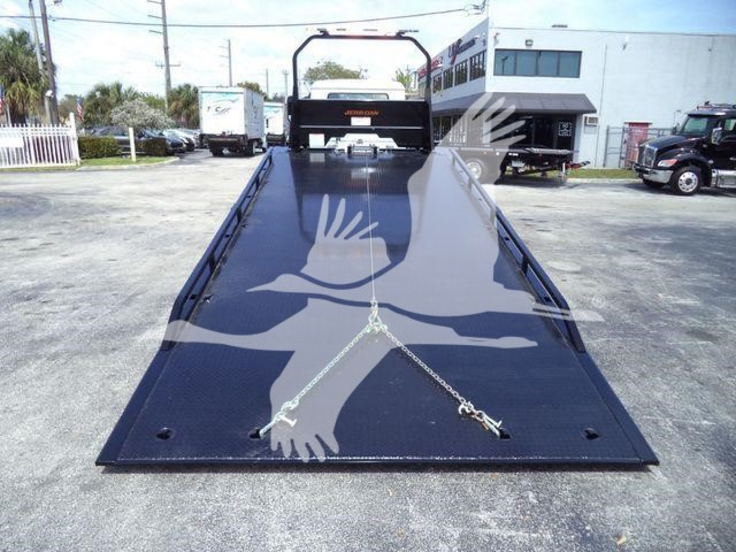 MD6 dump truck on flatbed rollback showing black steel deck and ramp with safety hooks, Mack branding at rear, surrounding lot with trucks.