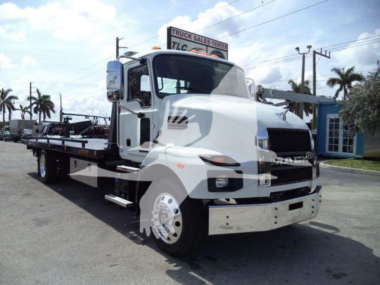 White Mack MD6 tow truck in a dealership lot, front three-quarter view, with flatbed and chrome bumper; TLC Truck Sales Center sign in background.