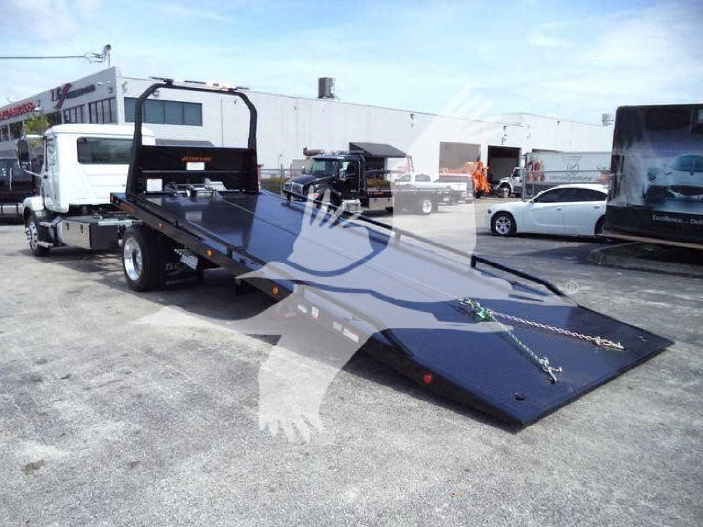 2027 Mack MD6 rollback truck with a large black flatbed/dropped ramp in foreground and a mounted gooseneck. visible text on shop and other vehicles in background.
