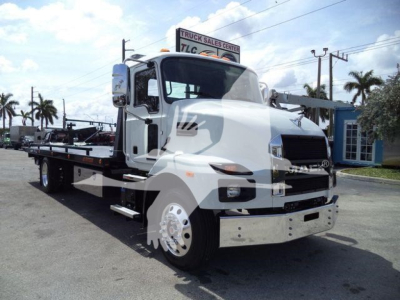 White Mack MD6 tow truck in a dealership lot, front three-quarter view, with flatbed and chrome bumper; TLC Truck Sales Center sign in background.