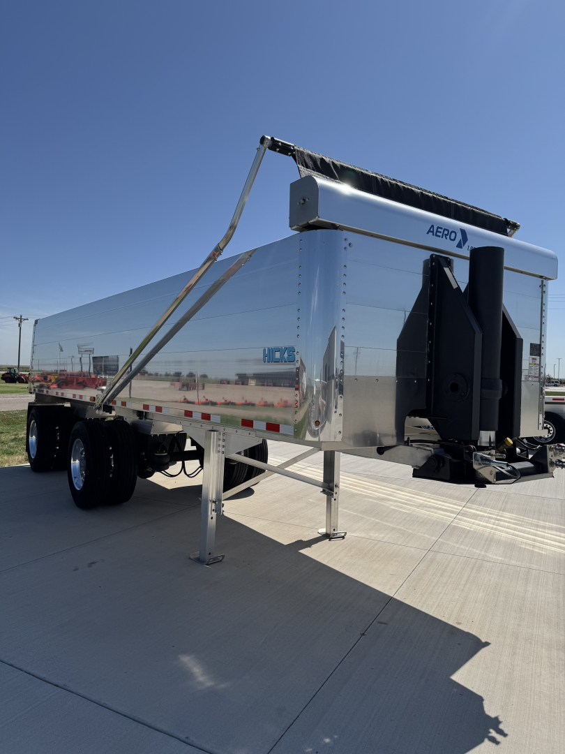 A chrome-dominant dump trailer with spread axle and hydraulic hoist, exterior chrome panels, tandem wheels, front hitch, and support leg visible.