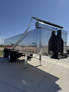 A chrome-dominant dump trailer with spread axle and hydraulic hoist, exterior chrome panels, tandem wheels, front hitch, and support leg visible.