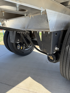 Trailer underside showing spread axle with metal deck and suspension arms, dual wheels, star shaped lug nuts, and hydraulic/air components visible.