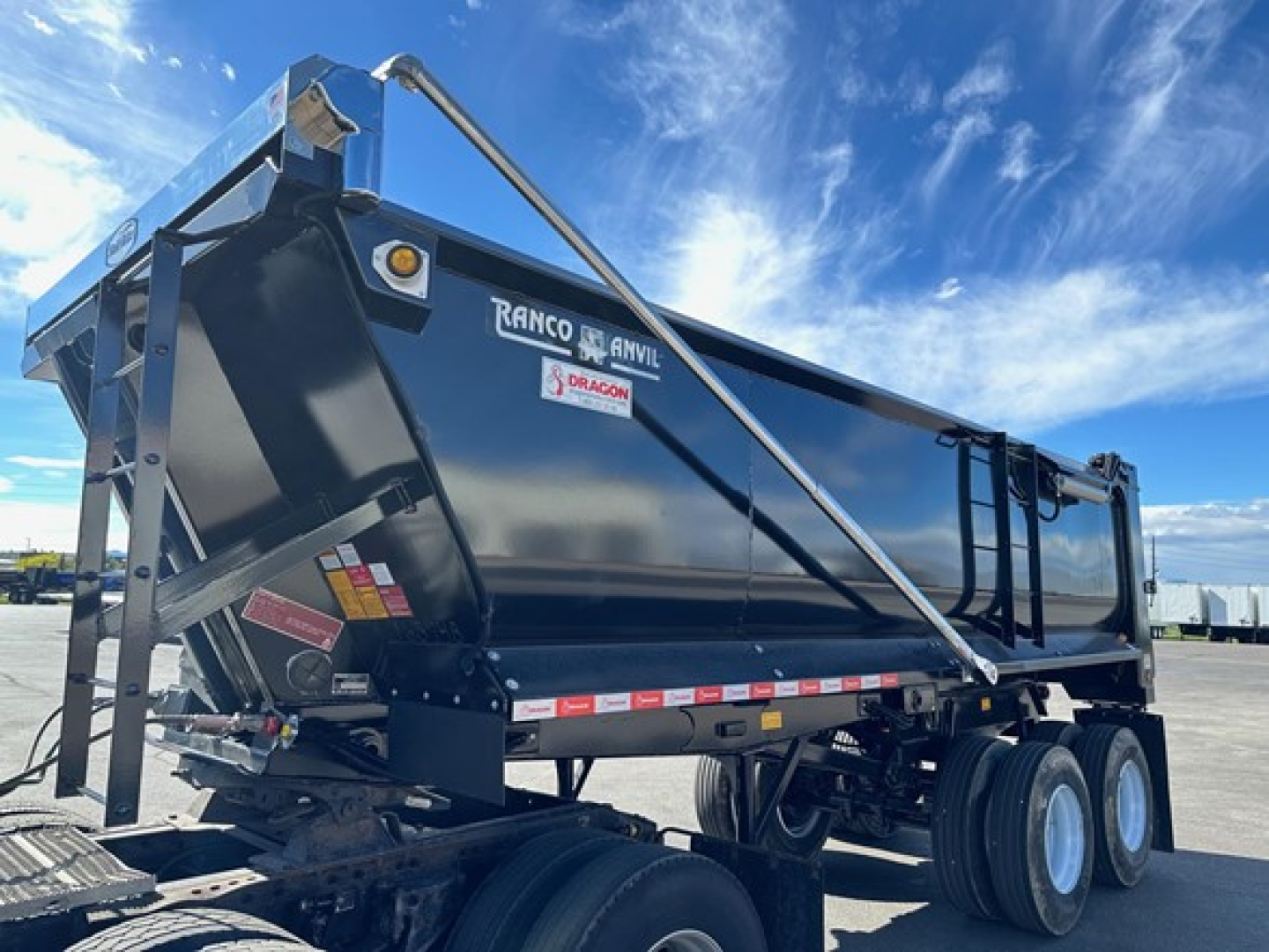 Ranco quarter frame steel end dump trailer with three-axle yard tires and hydraulic hoist, black finish, side decals and rear frame braces visible.