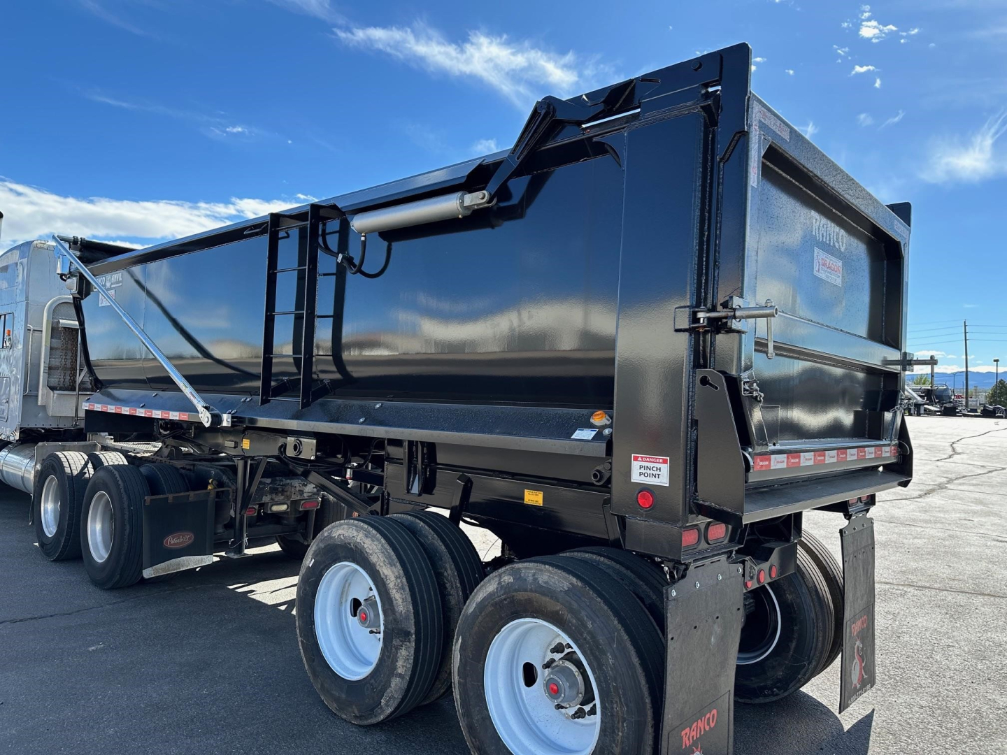 Ranco quarter-frame steel end dump trailer in black, three-axle with dual-tandem rear wheels, side panels, hydraulic hoist cylinder, and rear tailgate in open position.