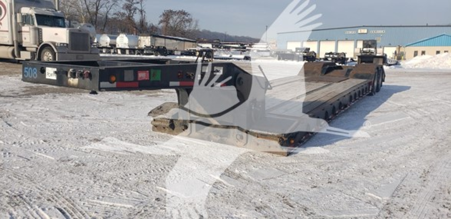 Lowboy trailer in a snowy lot showing beavertail section and multiple axles; no text legible except small numbers on rear fender.