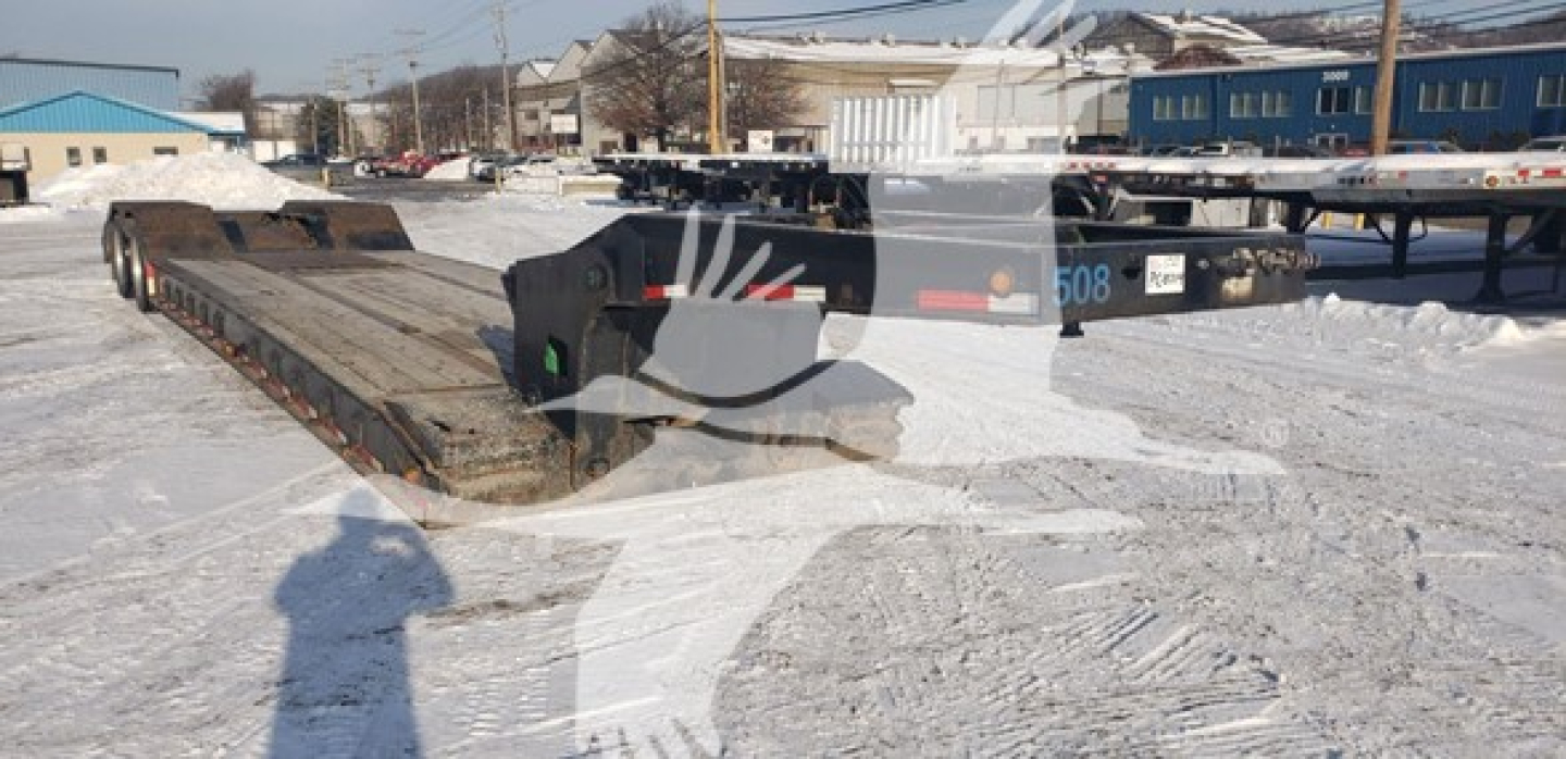 Lowboy trailer with beavertail showing flatbed deck and rear axles; there are numbers and labels on the hitch area blurred by shadow.